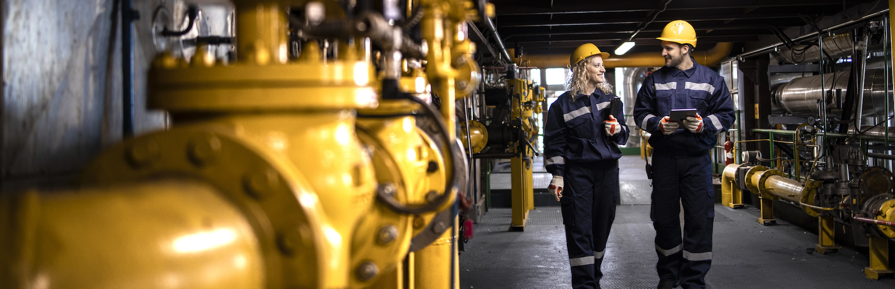 Two employees in yellow hard hats walking through an oil and gas facility 