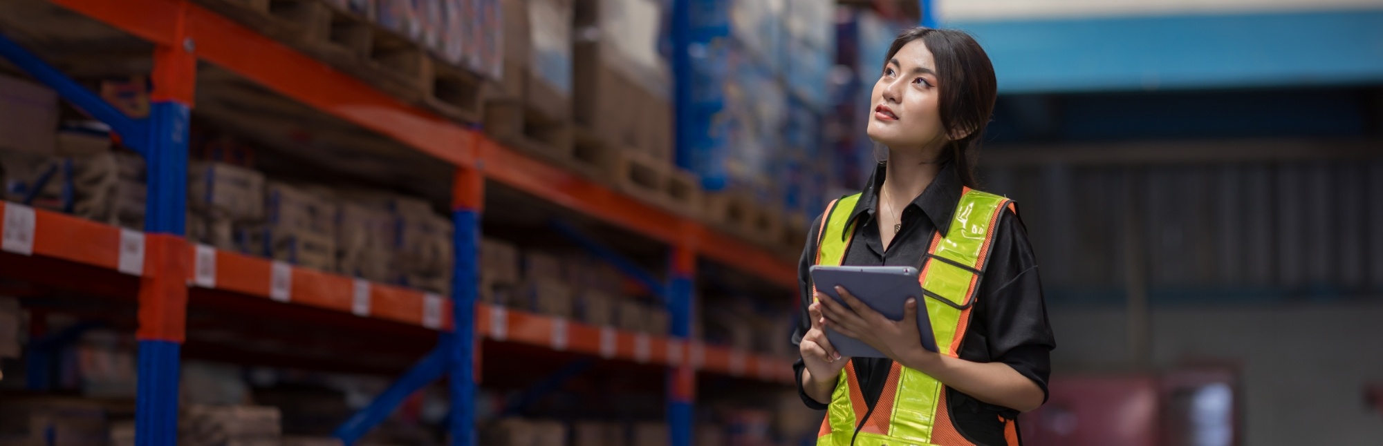 Woman in safety vest holding a tablet and looking up inside an industrial distribution center